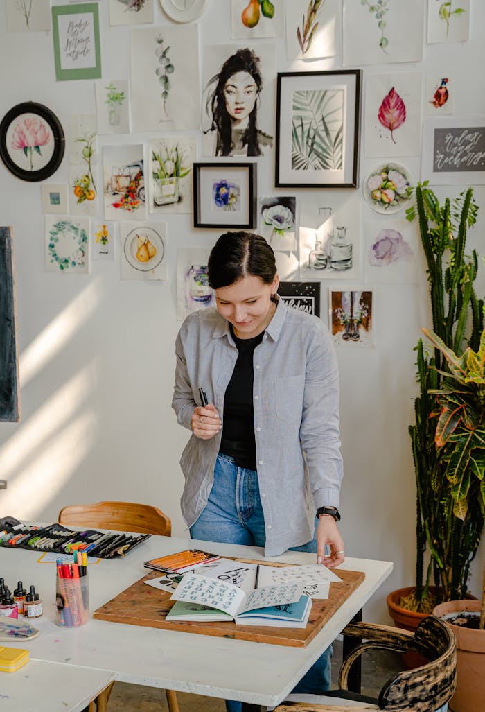 Cheerful young female designer in jeans standing near wall with pictures and table with stationery while choosing drafts with letter design in daylight